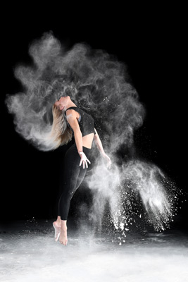 A black-clad dancer leaps into the air. A cloud of white dust spreads around her, emphasizing her movement.