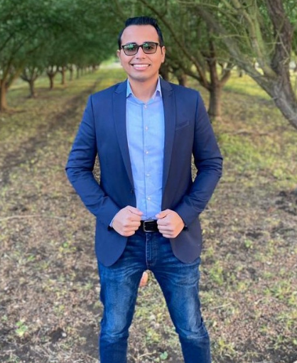 Jose is standing in the woods smiling with a blue jacket and jeans. 